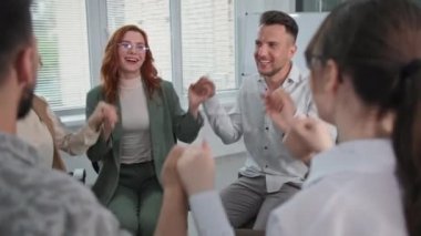 Çalışma takımı, neşeli erkek ve kadın meslektaşlar ofiste otururken el ele tutuşup alkışlıyorlar.