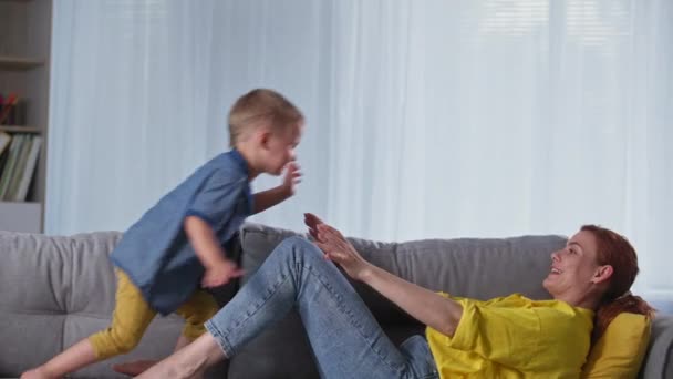 Vrolijke kindertijd, gelukkig jongetje heeft plezier met zijn moeder en springt naar haar voeten terwijl ontspannen op de bank thuis — Stockvideo