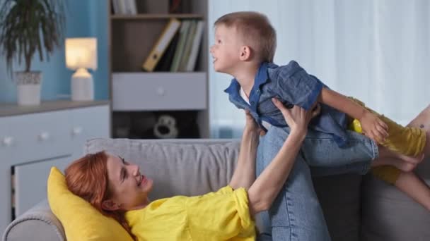 Macierzyństwo, szczęśliwa mama bawi się z dzieckiem i robi samolot leżąc na kanapie w pokoju — Wideo stockowe