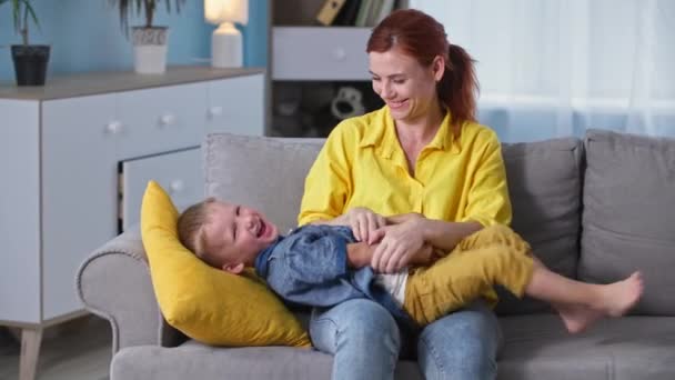 Nevelés, boldog fiatal anya élvezi a játékokat az ő kis örömteli fia, és csiklandozza őt, miközben otthon ül a kanapén — Stock videók