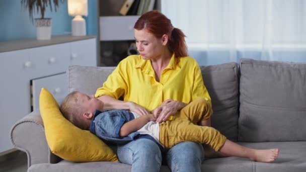 Happy male child has fun playing with his mom, loving female parent enjoys companionship with her son and tickles him sitting on couch in room — Stock Video