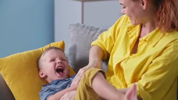 Boldog gondtalan anya és a kisfia szórakoznak társaságban, szerető női szülő gyengéden csiklandozza a férfi gyermek ül a kanapén otthon — Stock videók