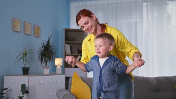 Rest at home, smiling young woman holding hands of cute male child actively jumping on trampoline in room — Stock Video