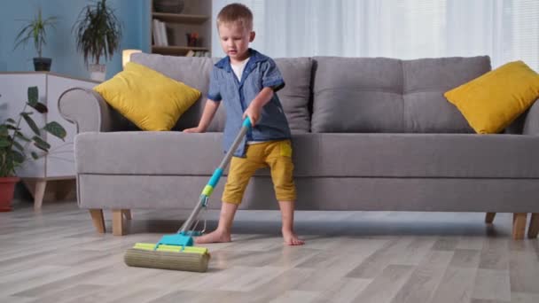Уборка дома, маленький трудолюбивый мальчик ребенок помогает родителям и моет пол с цветной шваброй в комнате — стоковое видео
