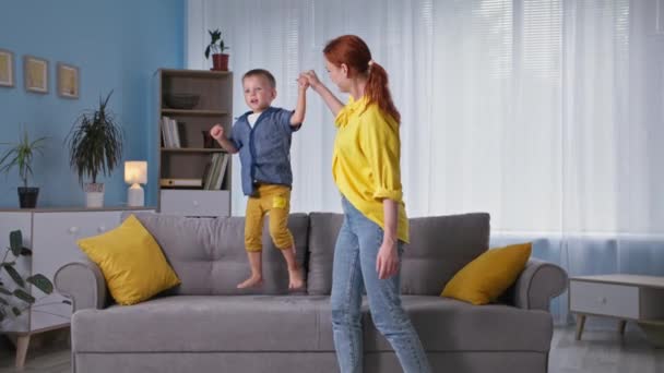 Adorable young female parent is having fun with their male child holding his hand as boy jumps on couch in room — Stock Video