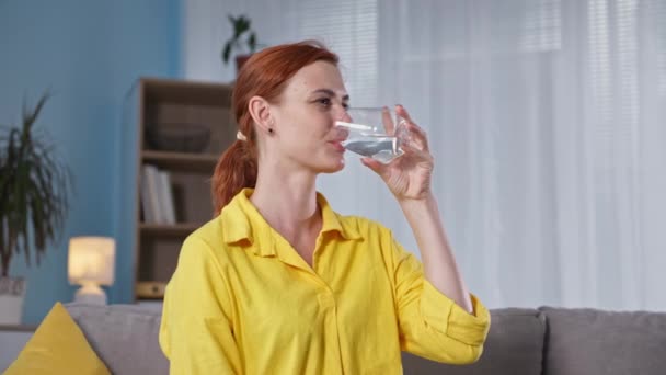 Portret van een jonge vrouw die haar dorst lessen en koud helder water drinken uit een glas terwijl ze op de bank in de kamer zit — Stockvideo