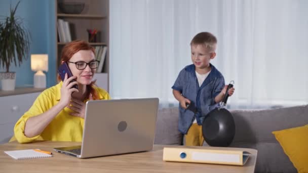 Escritório em casa, jovem mãe trabalha em casa e fala ao telefone enquanto seu filho interfere e brinca no sofá no quarto — Vídeo de Stock