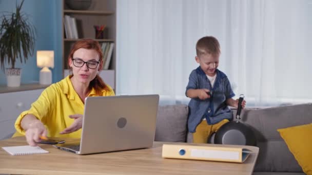 Bekerja pada cuti hamil, orang tua perempuan muda bekerja pada laptop sementara anak kecilnya membuat kebisingan dan mengganggunya sambil duduk di sofa di kamar — Stok Video