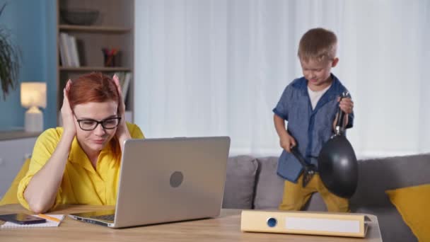 Nuori nainen työskentelee kannettavalla tietokoneella ja pitää päänsä, kun hänen miespuolinen lapsensa tekee melua ja leikkii istuessaan sohvalla huoneessa. — kuvapankkivideo