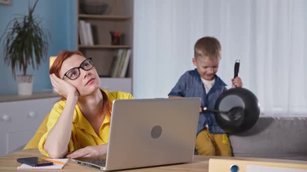 Freelancer, young mom works at laptop and holds her head while her male child makes noise and plays with frying pan while sitting on couch in room — Stock Video