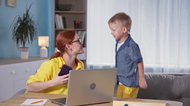 Jonge moeder is een zakenvrouw werken op laptop en praten over de telefoon terwijl haar zoontje springt op de bank in de kamer — Stockvideo