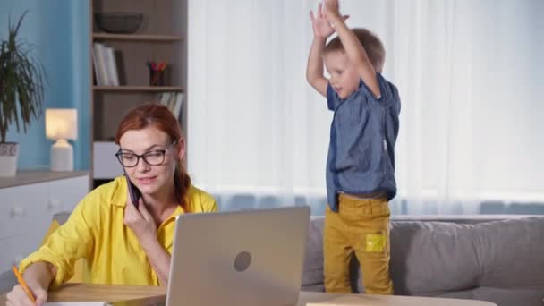 Ebeveynlik, iş kadınlığı dizüstü bilgisayarda çalışmak ve telefonla konuşmak. Bu arada küçük oğlu da oynuyor ve kanepede zıplayıp gürültü yapıyor. — Stok video