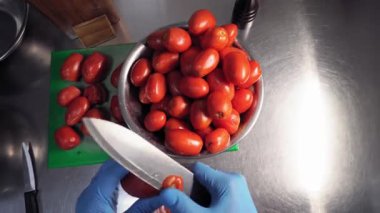 Mavi eldiven giyen şef, keskin bir mutfak bıçağıyla bir kaseden domatesleri alıyor, domateslerin kullanılmayan kısımlarını kesiyor ve onları yakınlardaki bir kesme tahtasına yerleştiriyor. Yukarıdan görünüm