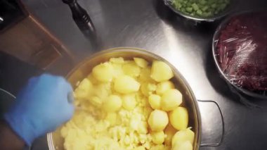 Mutfakta mavi eldiven takan bir aşçı kesim masasında haşlanmış patatesleri ezerek eziyor. Birinci şahıstan en iyi görüntü. Büyük yakın çekim.