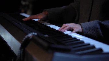 Düzgün manikürlü parmakları olan bir kadın elektrikli piyanoda sakin bir müzik çalar. Kadın, belirgin kumaş dokusuna sahip gri uzun kollu bir ceket giyiyor. Büyük yakın çekim.