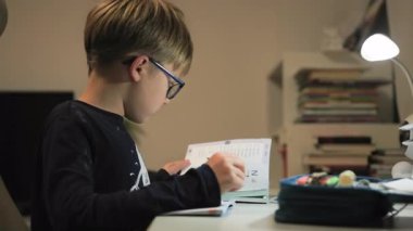 Gözlüklü bir çocuk masaya oturur ve defterine not alarak günlüğünü karıştırır. Yazı aletleri açık bir kalem kutusunun içinde masanın üzerinde yatıyor. Yakın çekim.