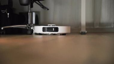Çalışan bir robot elektrikli süpürge temizlik yaparken mobilyaların etrafında dolaşır. Robot apartmanın ahşap zemininde hareket ediyor. Büyük yakın çekim. Yavaş çekim