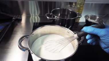 Mutfakta mavi eldiven giyen bir aşçı, ısıtmalı sütü ocakta çırpıcıyla karıştırıyor. Birinci şahıstan en iyi görüntü. Büyük yakın çekim.