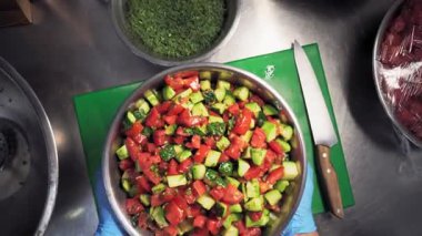 Aşçı mutfakta, kesim masasında mavi eldivenler giyerek salatayı salatalık, salatalık, domates ve dereyi büyük bir kasede karıştırmış olarak gösteriyor. Birinci şahıstan en iyi görüntü. Büyük yakın çekim.