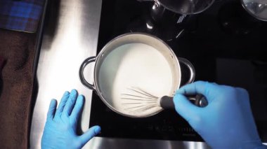 Mutfakta mavi eldiven giyen bir aşçı, ısıtmalı sütü ocakta çırpıcıyla karıştırıyor. Birinci şahıstan en iyi görüntü. Büyük yakın çekim.