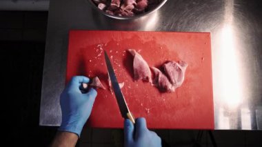 Şef, eti hızlıca ve dikkatlice kırmızı bir kesme tahtasının üzerinde küçük parçalara ayırmak için bir mutfak bıçağı kullanır, sonra onları yakındaki bir kaseye koyar. Birinci şahıstan en iyi görüntü. Büyük yakın çekim.