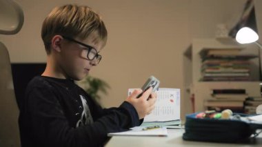 Gözlüklü bir çocuk odasında bir masada oturur ve bir deftere bir şeyler yazar. Sonra akıllı telefonunu aldı, bir mesaja cevap verdi ve telefonu yerine koydu. Yakın çekim.