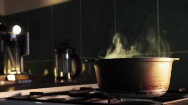 Karanlık bir mutfakta, bir adam elindeki makarnayı gaz sobasındaki kaynar su dolu bir kaba yavaşça döküyor. Mutfağın koyu arkaplanı tencerenin buharı ile keskin bir tezat oluşturuyor. Büyük yakın çekim.