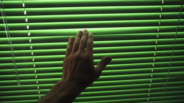 Pencere kenarında, bir adam sol elini aşağı hareket ettirir, parmaklarıyla kapalı yatay perdelere dokunur. Parmaklarının jaluziler boyunca hareketi ve dışarıdaki parlak güneş ışığı ince bir kurnazlık yaratır.