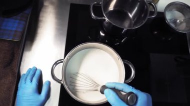 Mutfakta mavi eldiven giyen bir şef ısıtılmış sütü tencerede çırpıyla karıştırıyor. Sonra, başka bir fırına geçti, et sosunu başka bir tencerede kaşıkla karıştırdı. Üst görünüm