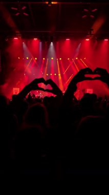 Hayranların karanlık silueti elleri kalp parmakları şeklinde, kırmızı arka plan gösterisinde, en sevdikleri rock grubu sahnede performans sergiliyor. Dikey video. Orta boy yakın çekim.
