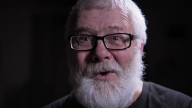 Gri saçlı gözlüklü adam dinliyor, sonra aniden şaşırıp sevinçle tepki veriyor, olanlar hakkında yüksek sesle yorum yapmaya devam ediyor. Koyu arkaplan, erkek figürüne şiddetle zıt düşüyor.