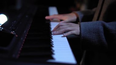 Düzgün manikürlü parmakları olan bir kadın elektrikli piyanoda sakin bir müzik çalar. Kadın, belirgin kumaş dokusuna sahip gri uzun kollu bir ceket giyiyor. Büyük yakın çekim.