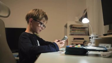 Bir çocuk ders sırasında masasında gizlice telefonunu ve sosyal medyasını kullanıyor. Akıllı telefonunu alır, bir mesaja cevap verir, ve annesi onu gördüğünde hemen yerine koyar.