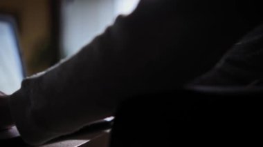 Genç adam evindeki kahve masasında klavye dizüstü bilgisayarında yazı yazıyor. Karanlık odada, pencereden gelen ışık klavyeye düşer ve adam ellerini tutar. Aşırı yakın çekim.