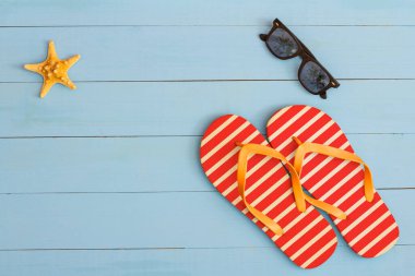 Yaz mevsimi, açık mavi ahşap bir masada güneş gözlüğü, parmak arası terlik ve deniz yıldızı ile serin bir manzara.
