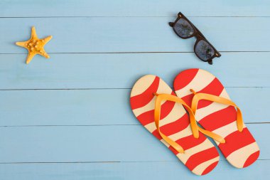 Yaz mevsimi, açık mavi ahşap bir masada güneş gözlüğü, parmak arası terlik ve deniz yıldızı ile serin bir manzara.