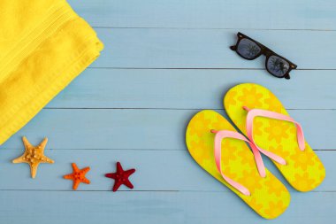 Yaz mevsimi, açık mavi ahşap masada güneş gözlüğü, terlik, havlu ve deniz yıldızı ile serin bir manzara.