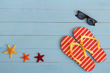Yaz mevsimi, açık mavi ahşap bir masada güneş gözlüğü, parmak arası terlik ve deniz yıldızı ile serin bir manzara.