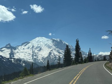 Glacier view framed by leafy trees in Mount Rainier National Park view from the windshield of a car with a long paved road.USA, Washington july 17 2025.