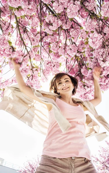 Young caucasian woman is jumping under the flowering sakura tree — Stock Photo, Image