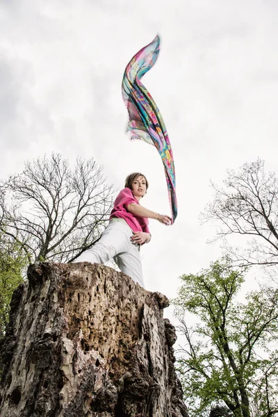 Молодая веселая женщина позирует на пне дерева с размахивающим шарфом — стоковое фото