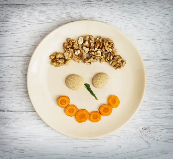 Cara divertida hecha de zanahoria, galletas y nueces en el plato — Foto de Stock