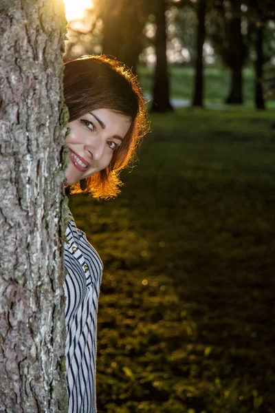 Young brunette woman is popping out behind the tree by sunset — Stock fotografie