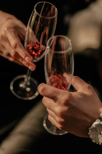 Miłość para trzyma pusty kieliszek szampana z czerwonym konfetti w kształcie serca. Miłosny eliksir w szkle. Zamknij się. — Zdjęcie stockowe