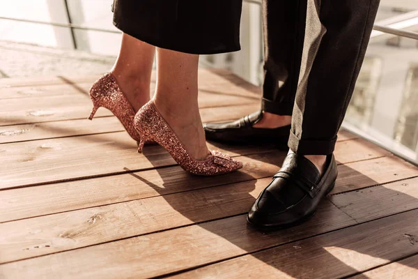 Acercamiento de una mujer en vestido negro elegante y rosa tacones altos de oro permaneciendo junto con su hombre en pantalones y zapatos negros. Elegante pareja que permanece frente a la luz del sol en el suelo de madera cerca de las barandillas — Foto de Stock