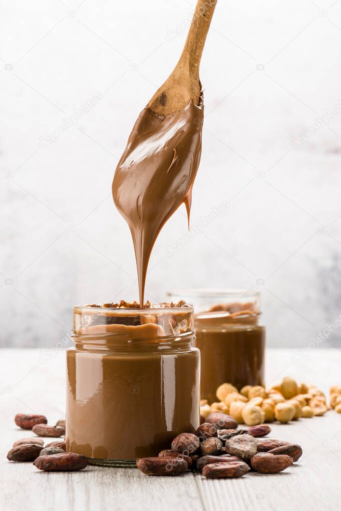 Chocolate cream in glass jar with cocoa beans, hazelnuts and dripping spoon, on wooden table