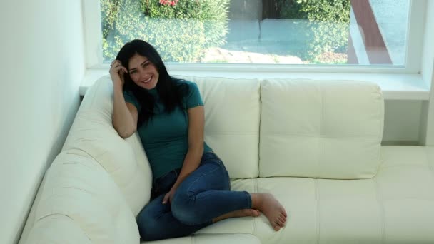 Linda mujer joven sentada en el sofá sonriendo mirando cámara — Vídeo de stock