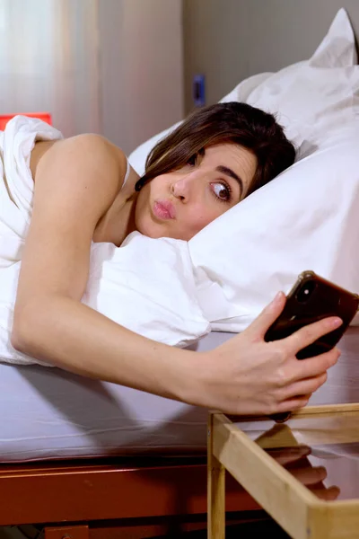 早上躺在床上用手机做鬼脸的有趣女人 免版税图库照片