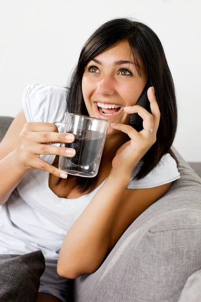 Leende yung kvinna prata i telefon medan du sitter på soffan dricka en kopp kaffe — Stockfoto