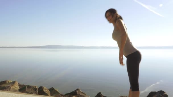Щаслива дівчина з придатним тілом робить розтягування перед озером під час красивого с — стокове відео